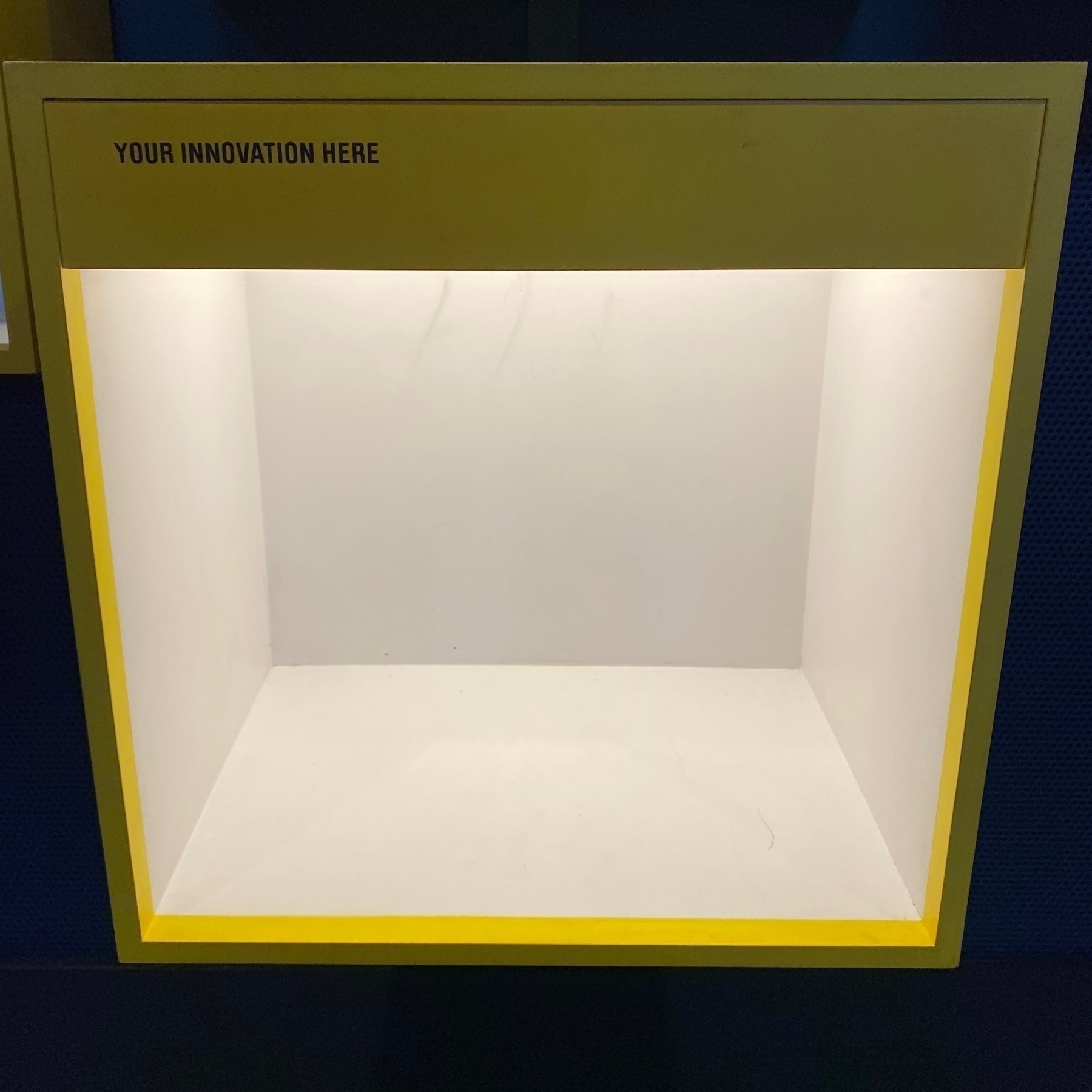 An empty museum display box titled 'Your Innovation Here', at the end of a wall of similar boxes filled with various inventions.