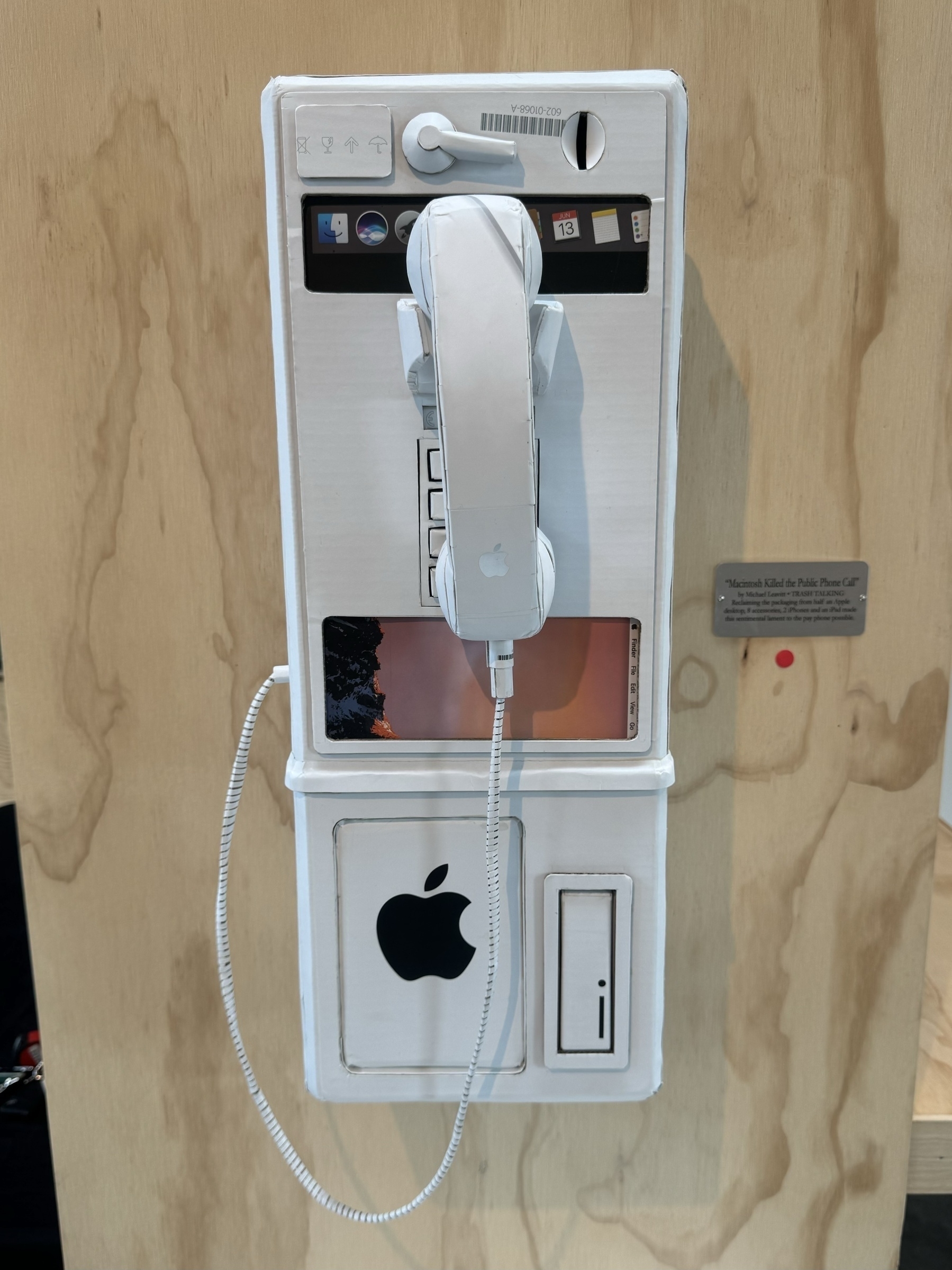 A art installation featuring a pay phone built entirely of used Apple packaging, mounted on a wooden backdrop.