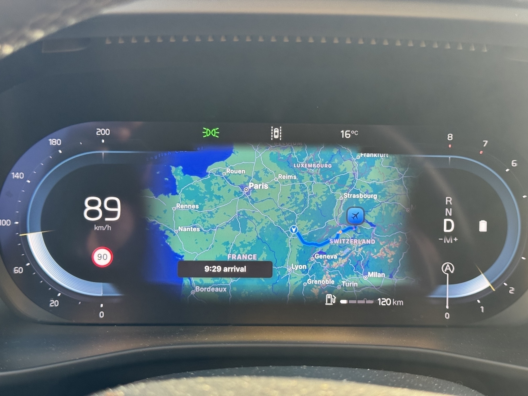 A photograph of a car dashboard display shows a navigation map with an arrival time, surrounded by speed, gear, and temperature indicators.