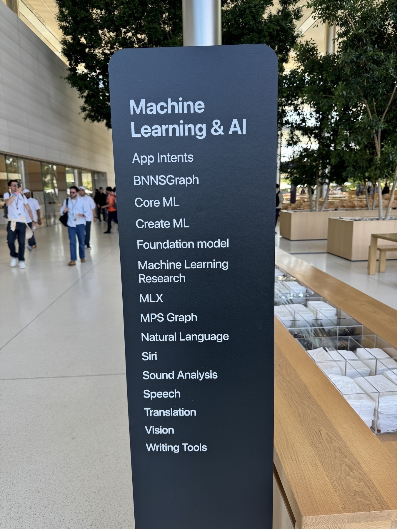 A sign inside Caffè Macs at Apple Park listing Machine Learning & AI topics including App Intents, BNNSGraph, Core ML, Create ML, Foundation model, ML Research, MLX, MPS Graph, Natural Language, Siri, Sound Analysis, Speech, Translation, Vision and Writing Tools. Indoor trees in the background. 
